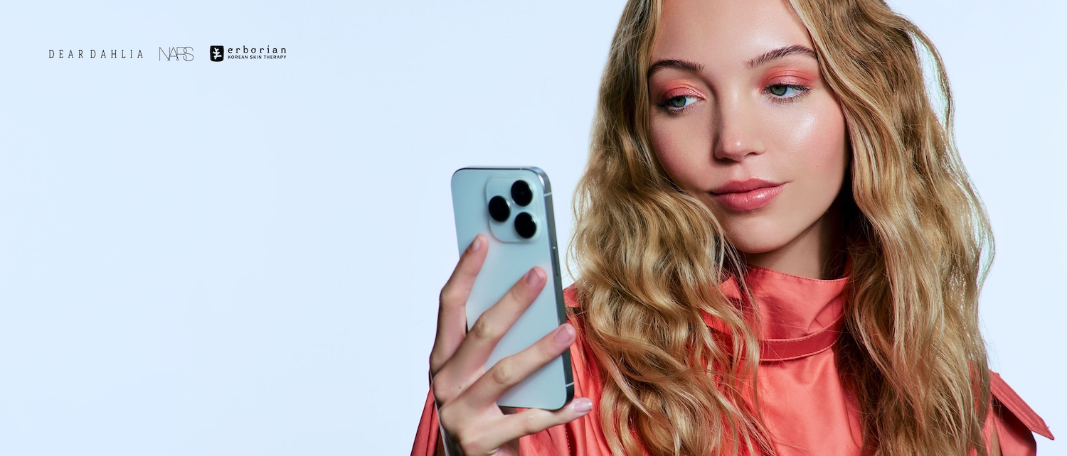 Una donna bionda con ombretto color pesca e labbra lucide tiene in mano un telefono cellulare; in alto a sinistra sono visibili i loghi di Dear Dahlia, NARS ed Erborian.