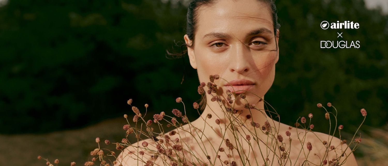 Primo piano di una modella con trucco naturale, che tiene dei fiori secchi davanti al petto, con il logo di Airlite x Douglas.