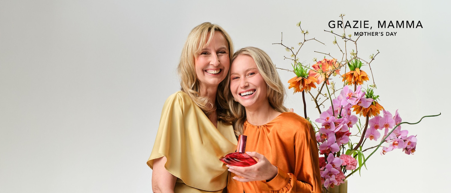 Festeggia la Festa della Mamma con un profumo in edizione limitata, presentato da due donne sorridenti in abiti color oro e arancio, accanto a un bouquet di fiori colorati.