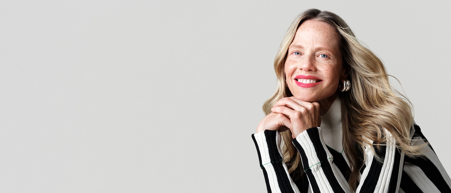 Una donna sorridente con capelli biondi, lentiggini e labbra rosa acceso, indossa un maglione a righe bianche e nere e orecchini a cerchio argentati.