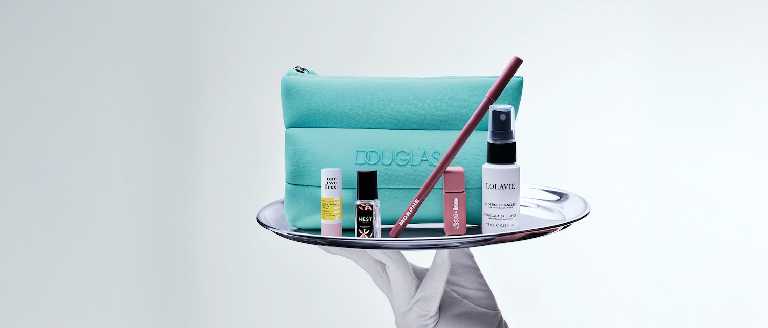 Un guanto bianco tiene un vassoio d'argento con una pochette turchese Douglas e prodotti di bellezza come rossetti, profumi e spray districante Lolavie.