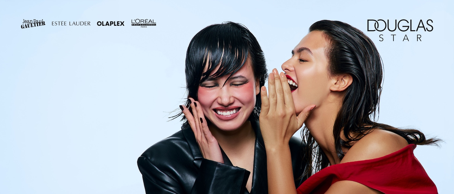 Due donne sorridono, una sussurra un segreto all'orecchio dell'altra, con loghi di marchi di bellezza come Jean Paul Gaultier, Estée Lauder, Olaplex e L'Oréal sullo sfondo e il logo Douglas Star.