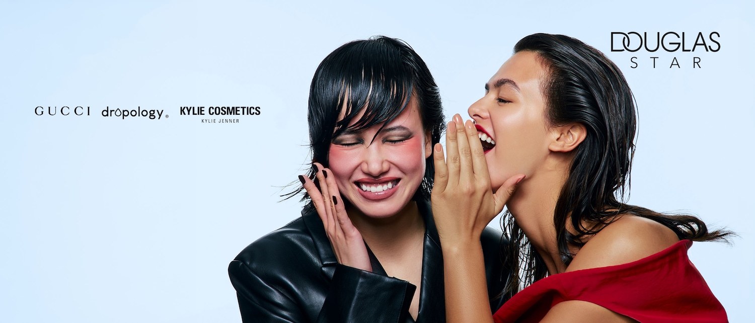 Due donne sorridono, una sussurra un segreto all'orecchio dell'altra, con loghi di marchi come Gucci, Kylie Cosmetics e Douglas Star.
