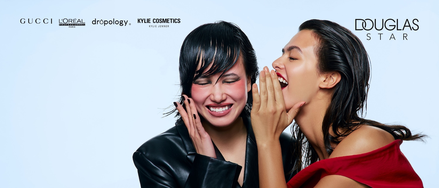 Due donne sorridono, una sussurra un segreto all'orecchio dell'altra, con loghi di marchi di bellezza come Gucci, L'Oréal, Kylie Cosmetics e Douglas Star.