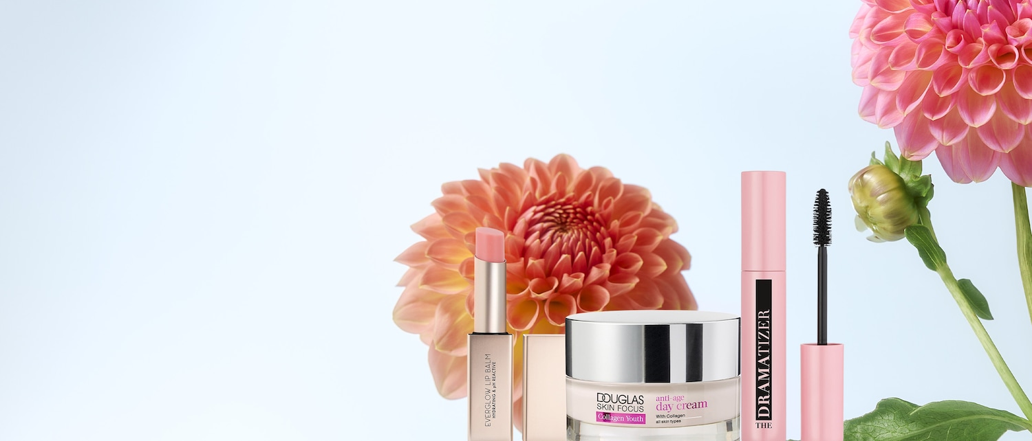 Ecco un balsamo labbra Everglow, un trattamento giorno anti-età Douglas Skin Focus Collagen Youth e un mascara Dramatizer, presentati su uno sfondo chiaro con fiori di dalia rosa e arancioni.