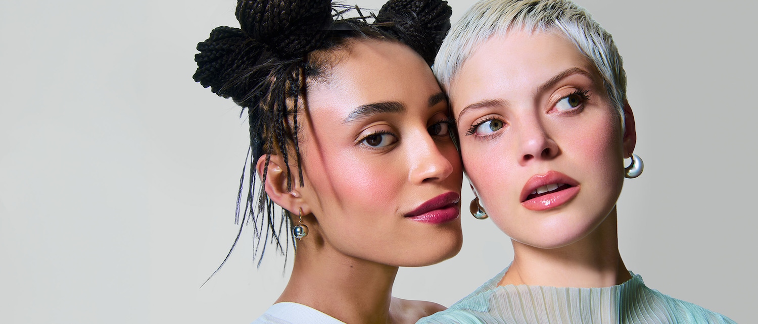 Primo piano di due donne con trucco luminoso e orecchini pendenti, una con trecce afro e l'altra con capelli biondo platino corti.