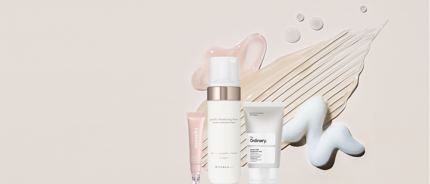 Una schiuma detergente delicata di Rituals, un burro labbra di Kylie Skin e una sospensione di acido azelaico di The Ordinary sono esposti su uno sfondo beige con schizzi di prodotto.