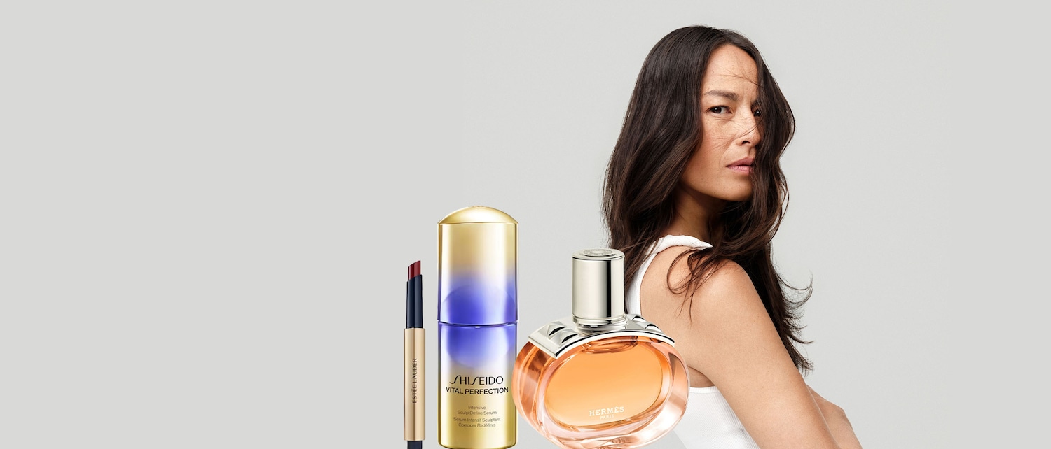 Una donna con lunghi capelli castani guarda oltre la spalla, con un rossetto Estée Lauder, un siero Shiseido Vital Perfection e un profumo Hermès in primo piano.
