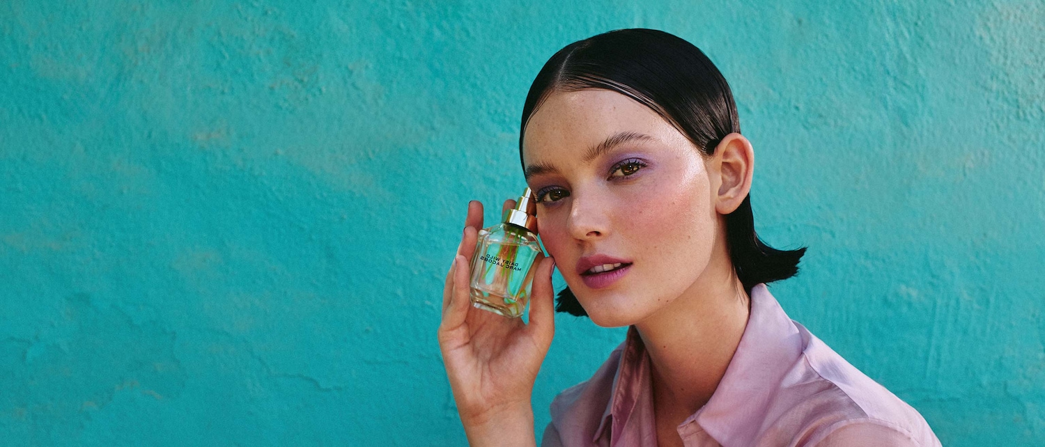 Una modella con ombretto viola e camicia rosa tiene in mano una bottiglia di profumo trasparente Marc Jacobs Daisy Eau So Fresh.