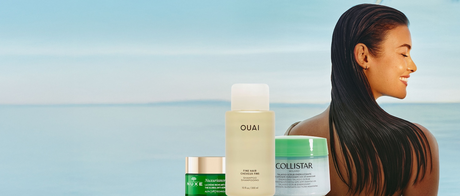 Primo piano di prodotti di bellezza Nuxe verde, Ouai color crema e Collistar bianco-verde, con una donna sorridente sullo sfondo, che mostra una gamma di prodotti per la cura della pelle e dei capelli.