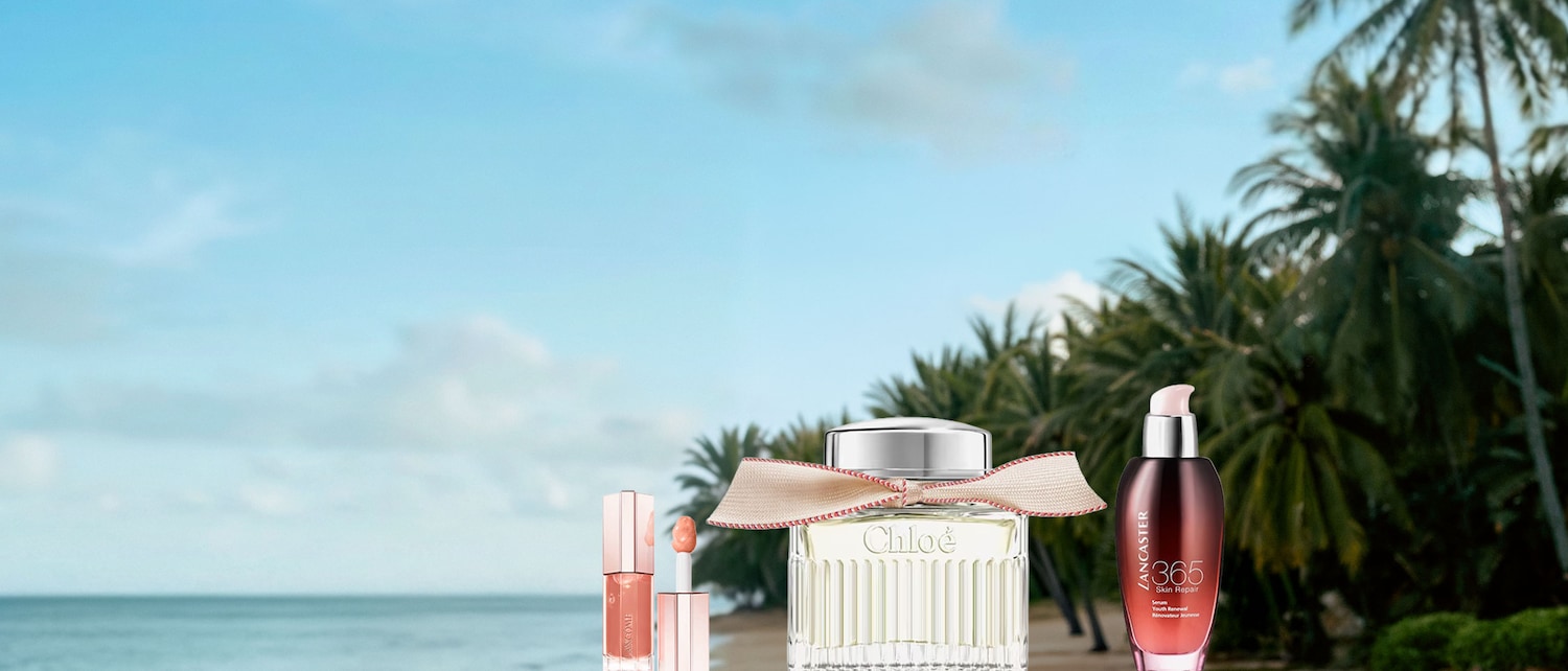 Sullo sfondo di una spiaggia tropicale, spiccano un profumo Chloé con nastro beige, un lucidalabbra Lancôme rosa e un siero Lancaster 365 Skin Repair.