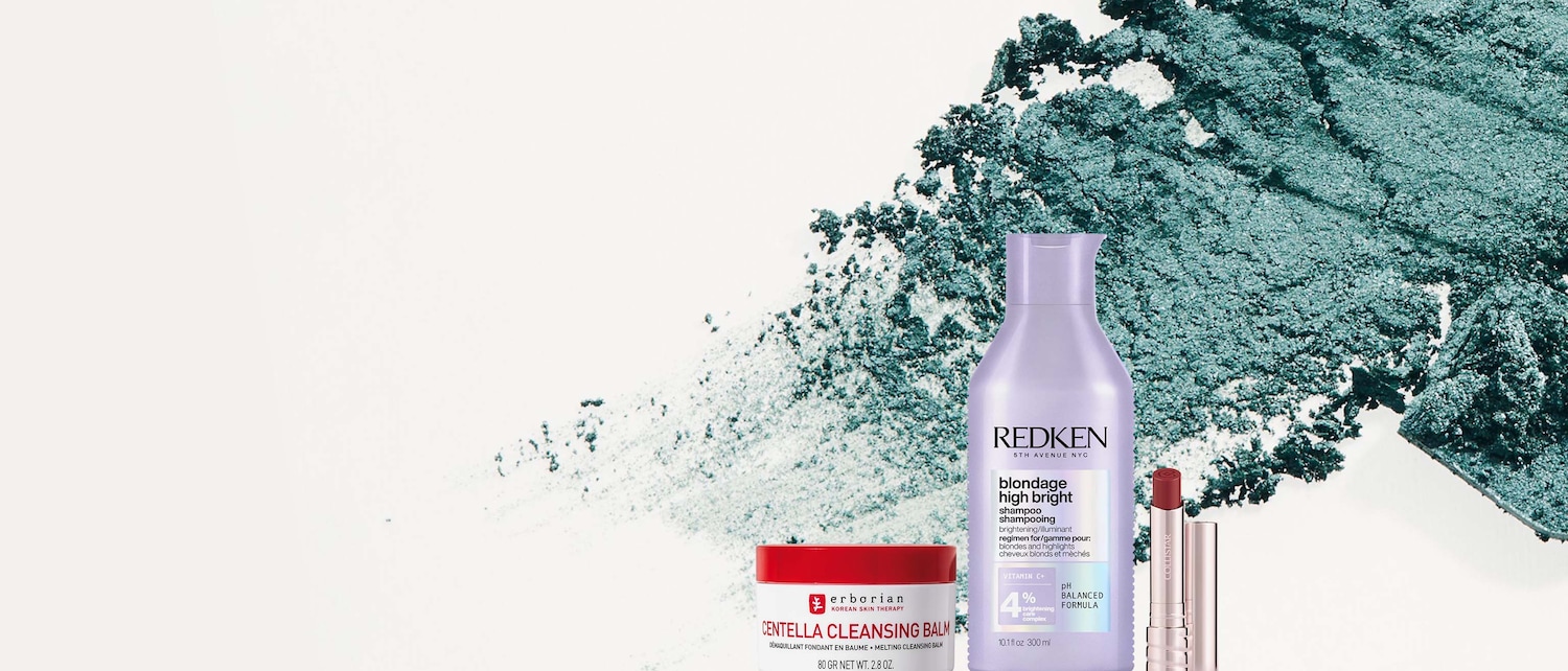 Un balsamo detergente Erborian Centella in un barattolo rosso e bianco, uno shampoo Redken Blondage High Bright in una bottiglia viola e due rossetti Collistar in tonalità rosse, tutti su uno sfondo bianco con spruzzi di polvere verde acqua.