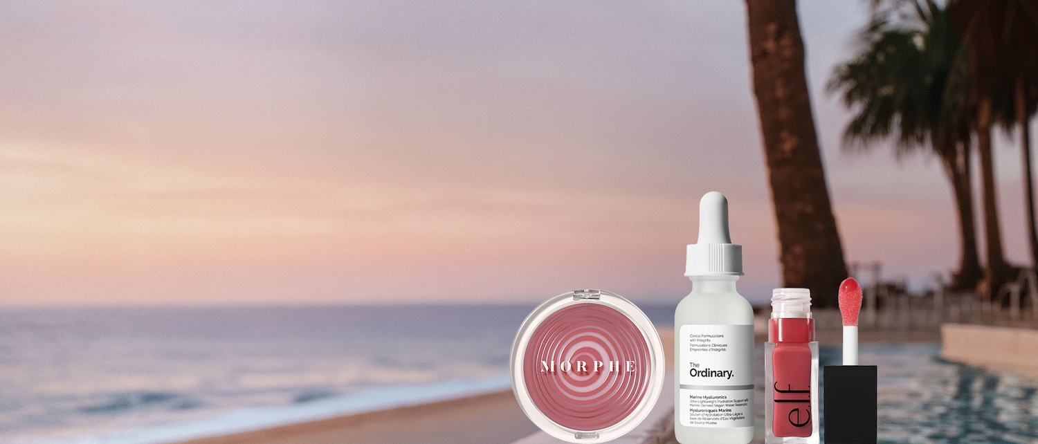 Un blush rosa Morphe, un siero The Ordinary Marine Hyaluronics e un lucidalabbra rosso Elf sono esposti su un parapetto con vista su un tramonto sull'oceano e palme.