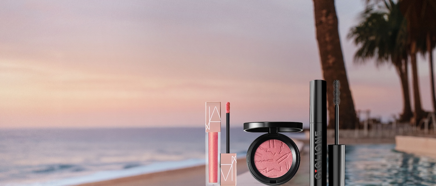 Un lucidalabbra rosa pesca con applicatore, un blush rosa chiaro con il logo MAC e un mascara nero con la scritta "CIGLIONE" sono esposti su uno sfondo sfocato di un tramonto sulla spiaggia con palme e una piscina.