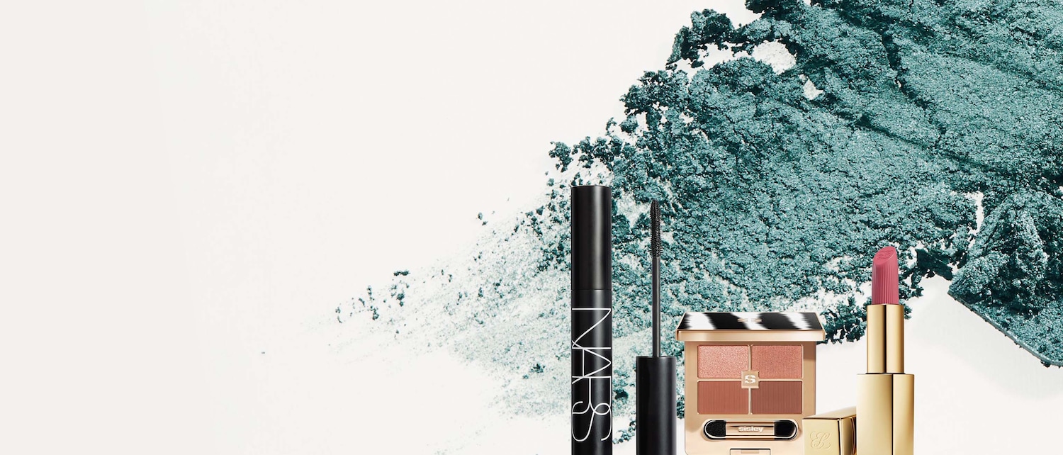 Un mascara nero NARS, una palette ombretti Sisley con quattro tonalità marroni e un rossetto rosa in un astuccio dorato sono disposti su uno sfondo bianco con una spolverata di ombretto verde acqua.
