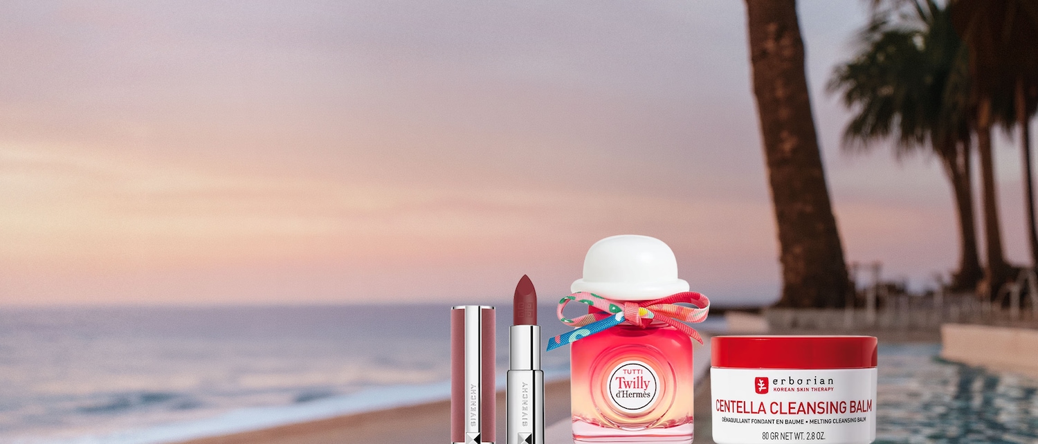 Due rossetti Givenchy, uno rosa chiaro e uno rosso scuro, sono affiancati a un profumo Twilly d'Hermès con un fiocco colorato e un balsamo detergente Erborian Centella, tutti posizionati su un bordo piscina con vista mare al tramonto.