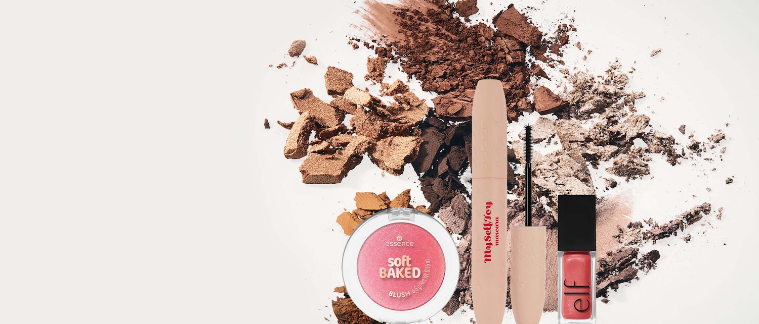 Un blush rosa chiaro "soft BAKED" di Essence, un mascara "My Self Toy" color pesca e un lucidalabbra rosso corallo di Elf sono disposti su uno sfondo bianco con ombretti in polvere marroni e bronzo sparsi.