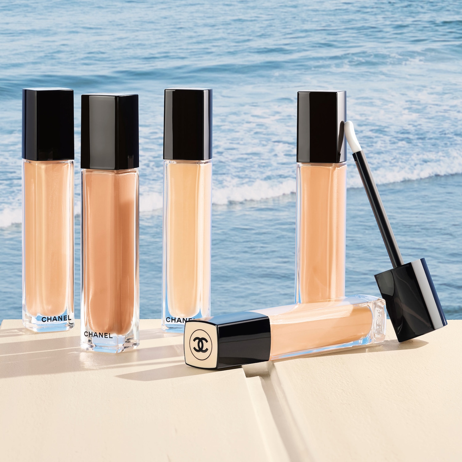Una fila di flaconi di fondotinta Chanel color pesca chiaro con tappi neri lucidi su una superficie beige, con l'oceano sullo sfondo.