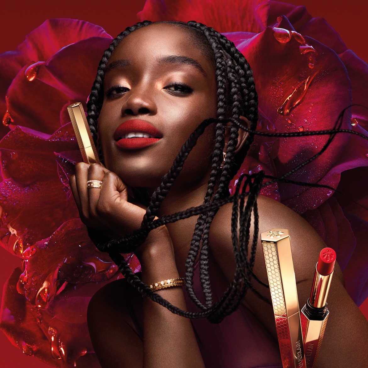 Primo piano di una modella con trecce che indossa un rossetto rosso opaco, con una rosa rossa sullo sfondo e un rossetto Guerlain dorato con motivo a nido d'ape in primo piano.