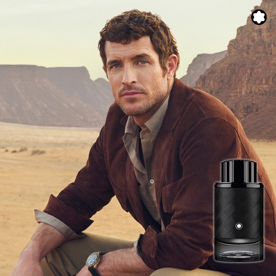 Un uomo con giacca marrone e orologio è seduto nel deserto con sullo sfondo montagne e una bottiglia di profumo Montblanc.