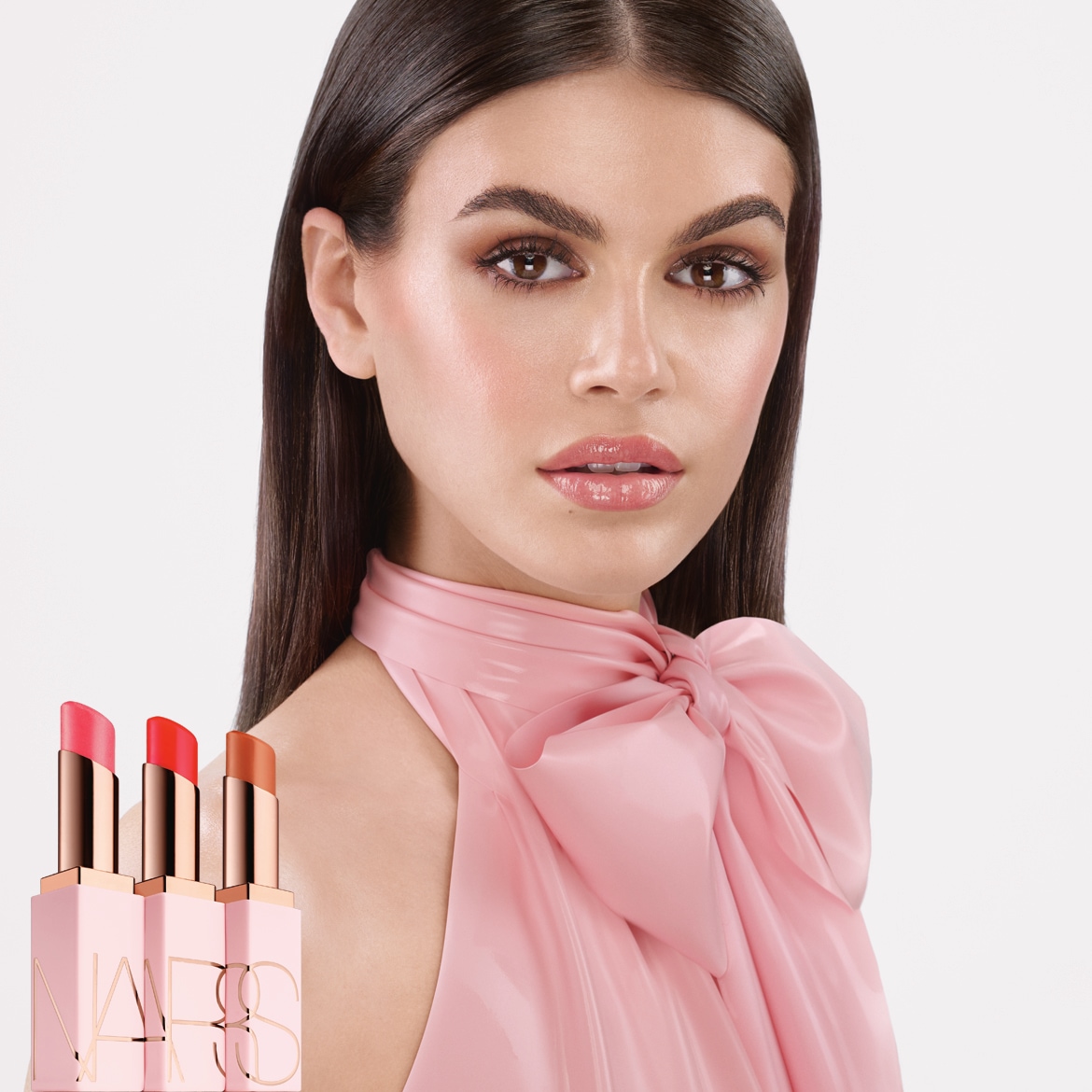 Primo piano di una modella con un abito rosa e tre rossetti NARS rosa con tonalità corallo, arancio e marrone, per labbra idratate e colorate.