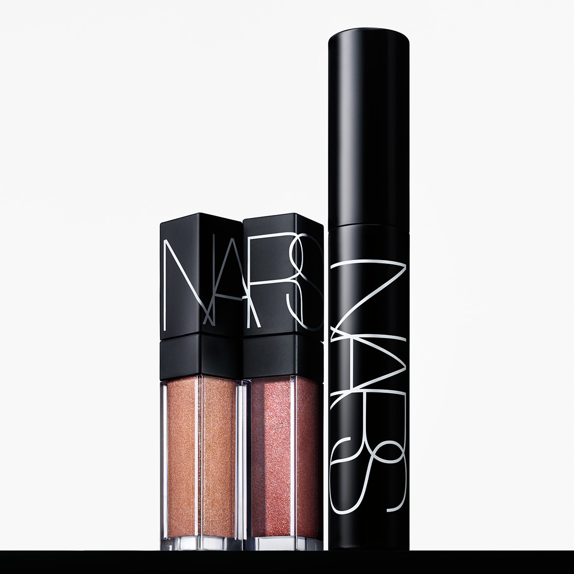 Due lucidalabbra NARS in tonalità oro e rosa scintillanti, affiancati da un mascara nero NARS, tutti con il logo bianco del marchio.