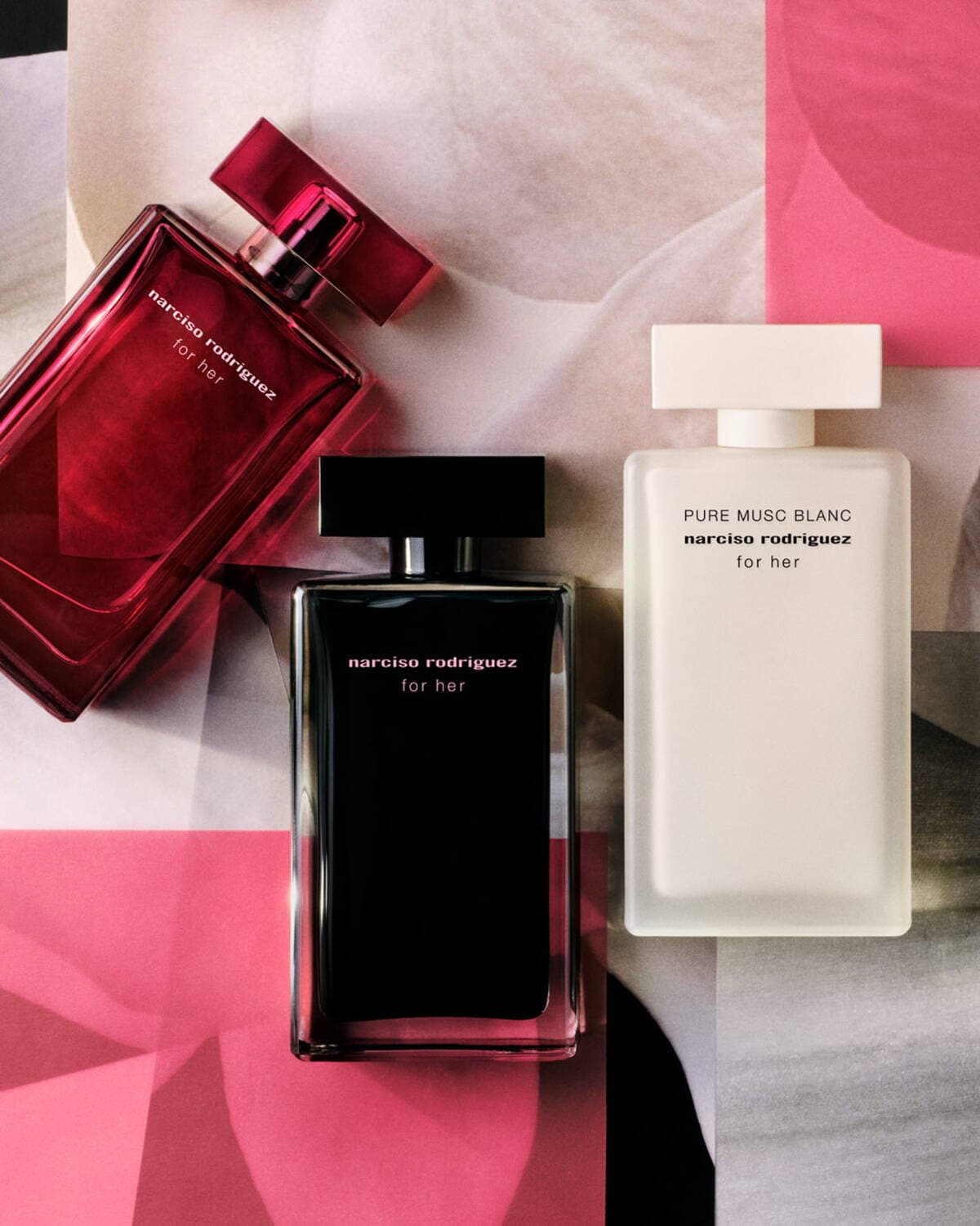 Tre flaconi di profumo Narciso Rodriguez "for her", uno rosso rubino, uno nero intenso e uno bianco puro, disposti su uno sfondo astratto nei toni del rosa e del beige.