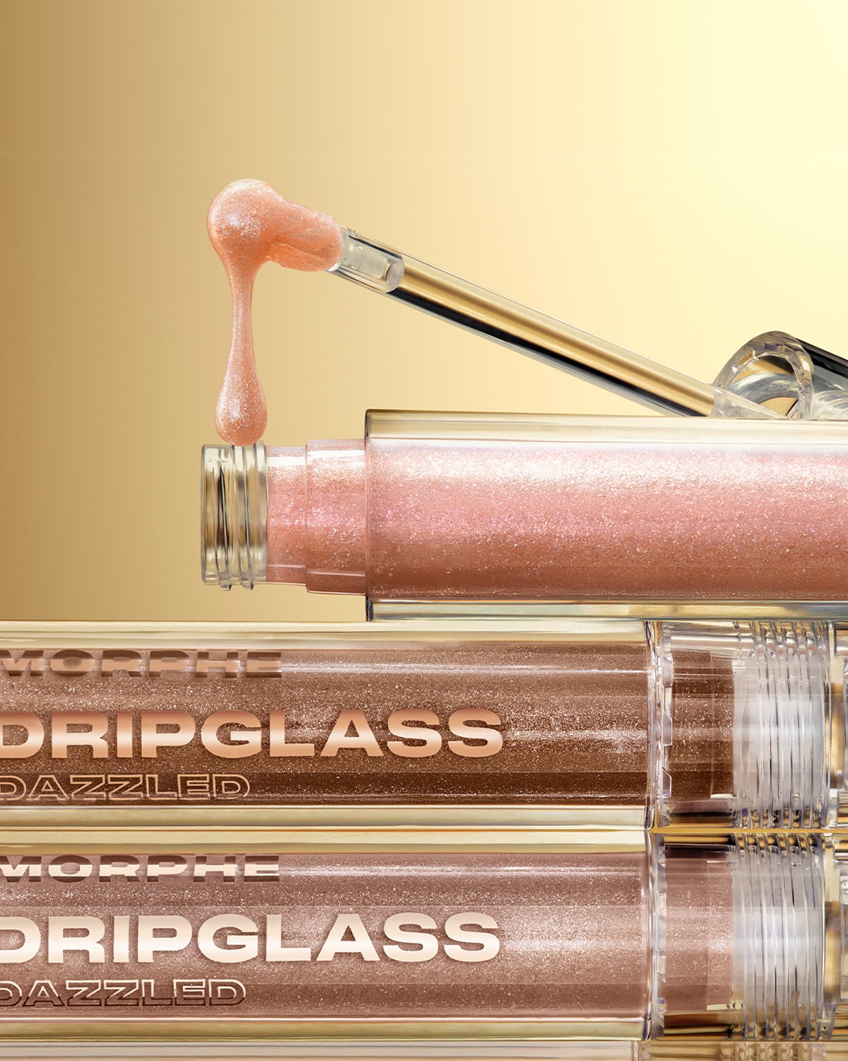 Ecco il lucidalabbra Dripglass Dazzled di Morphe con un applicatore trasparente che mostra una goccia di lucidalabbra rosa brillante, il tutto su uno sfondo dorato.