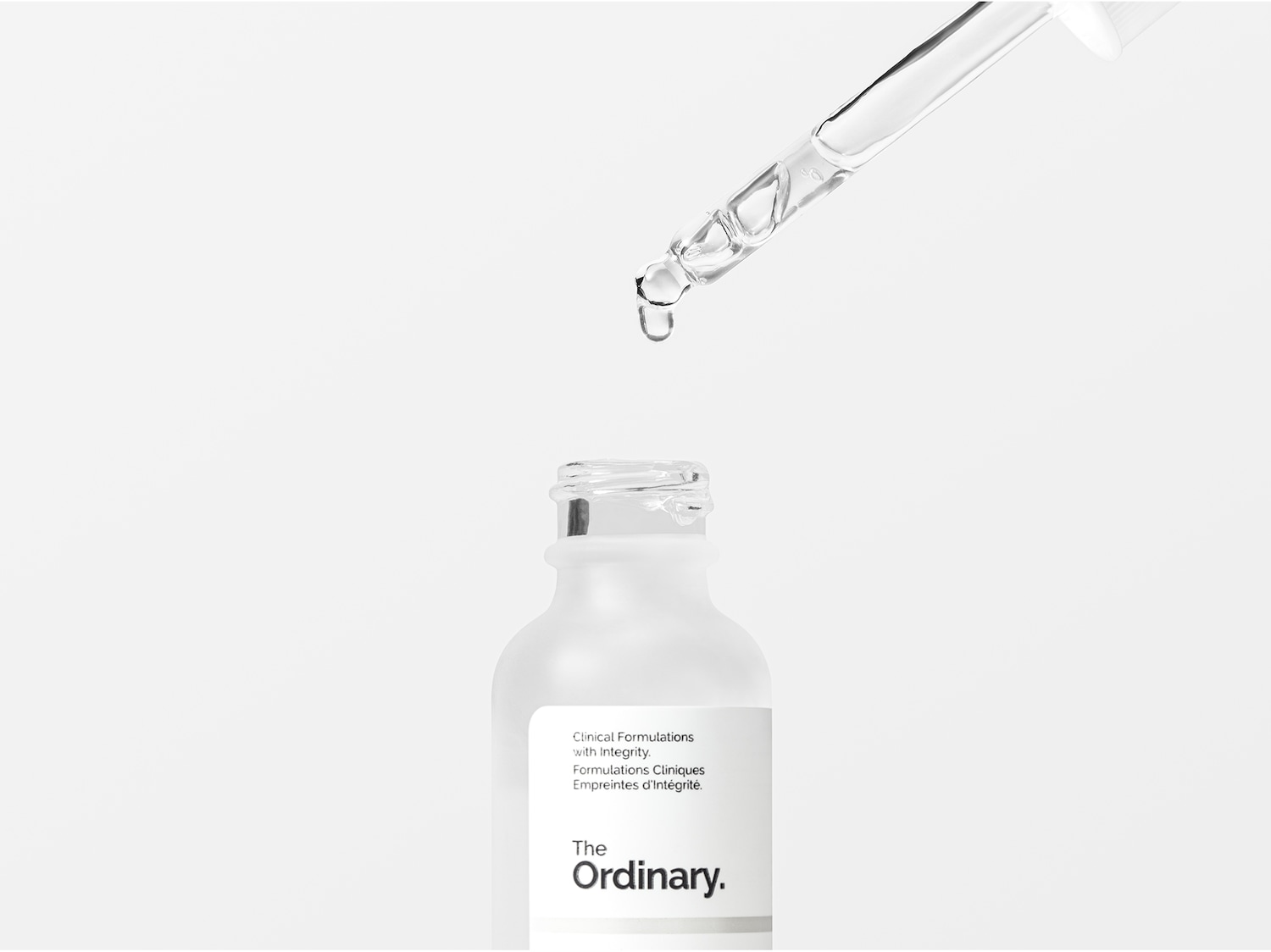 Ecco un flacone bianco opaco di The Ordinary con un contagocce trasparente che rilascia una goccia di siero, mettendo in risalto le formulazioni cliniche con integrità.