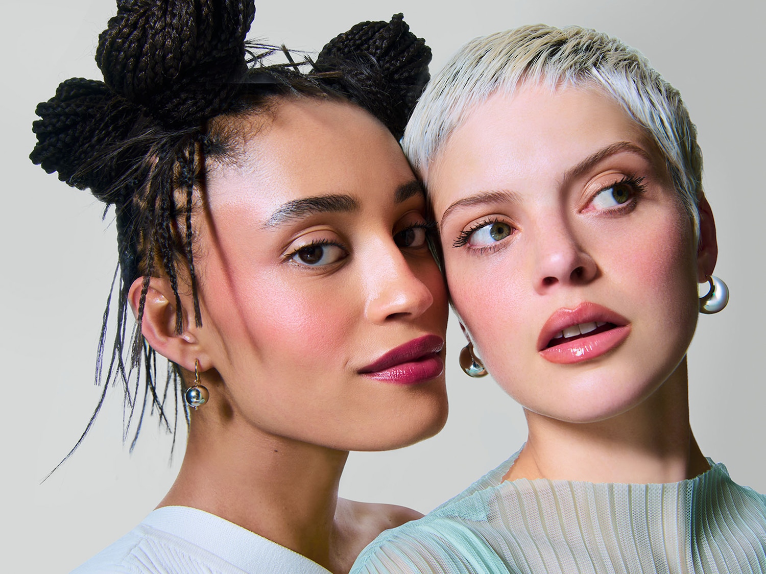 Primo piano di due donne con incarnato diverso, la prima ha i capelli neri raccolti in tre chignon e indossa orecchini dorati, la seconda ha i capelli biondo platino corti e orecchini argentati.