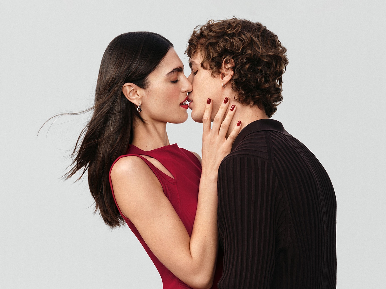 Primo piano di una coppia che si bacia appassionatamente, lei indossa un abito rosso e lui una maglia marrone a coste, entrambi con smalto rosso sulle unghie.