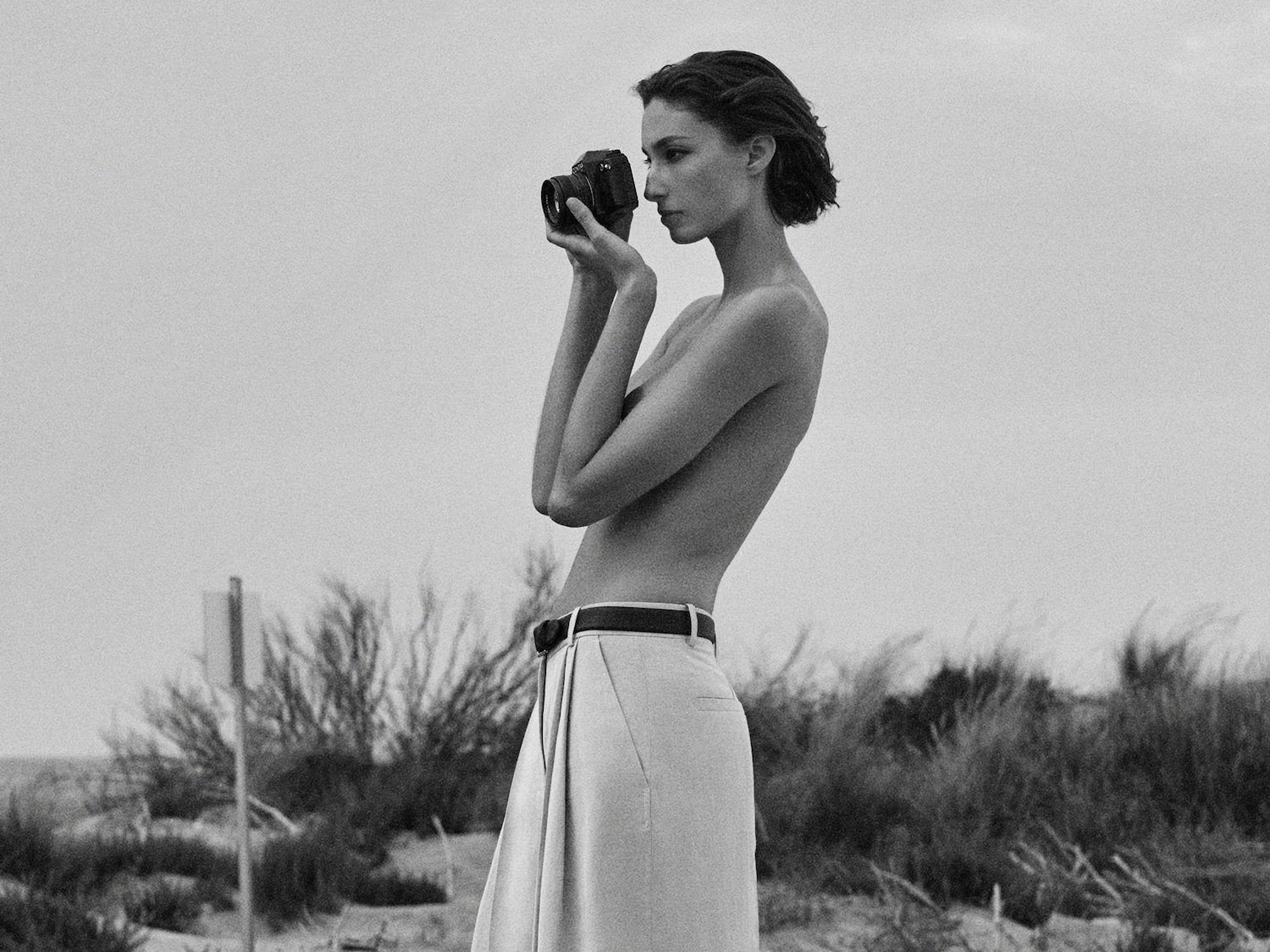 In una foto in bianco e nero, una donna con i capelli corti tiene in mano una fotocamera, indossando pantaloni chiari con una cintura scura, in piedi in un paesaggio naturale.