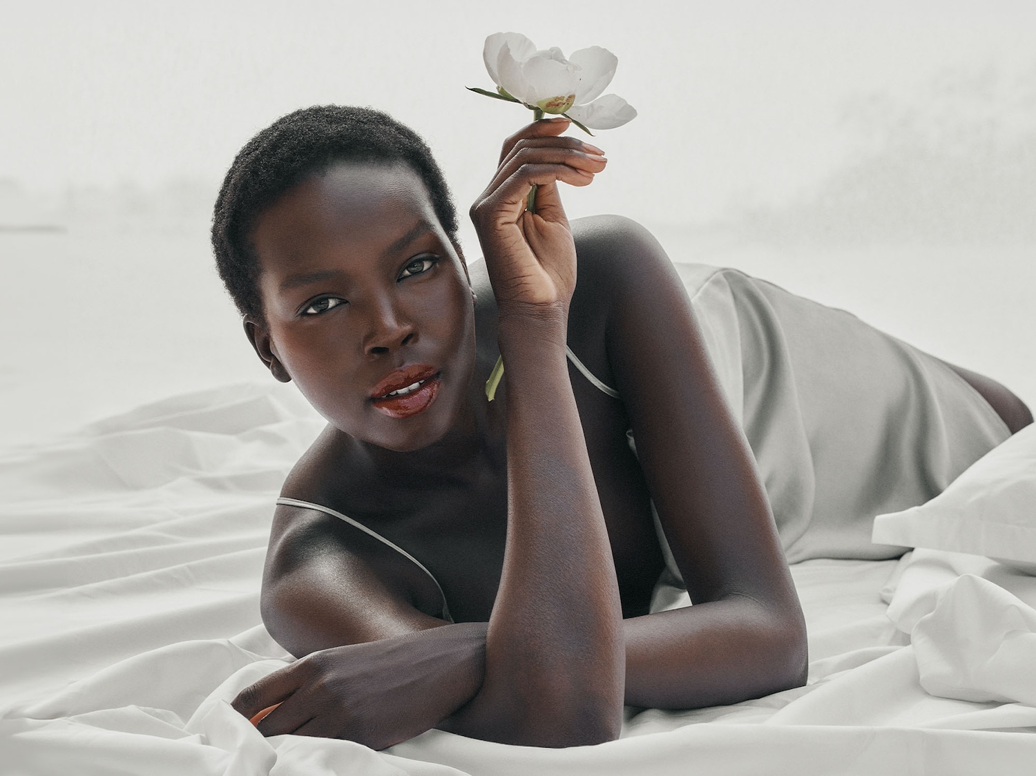 Una donna di colore con i capelli corti è sdraiata su un lenzuolo bianco e tiene in mano un fiore bianco, con un'espressione serena.