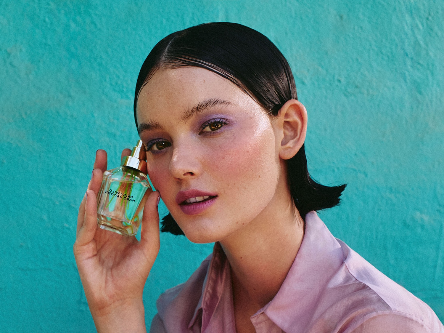 Primo piano di una donna con ombretto viola e labbra rosa, che tiene in mano una bottiglia di profumo trasparente con tappo dorato, su sfondo turchese.