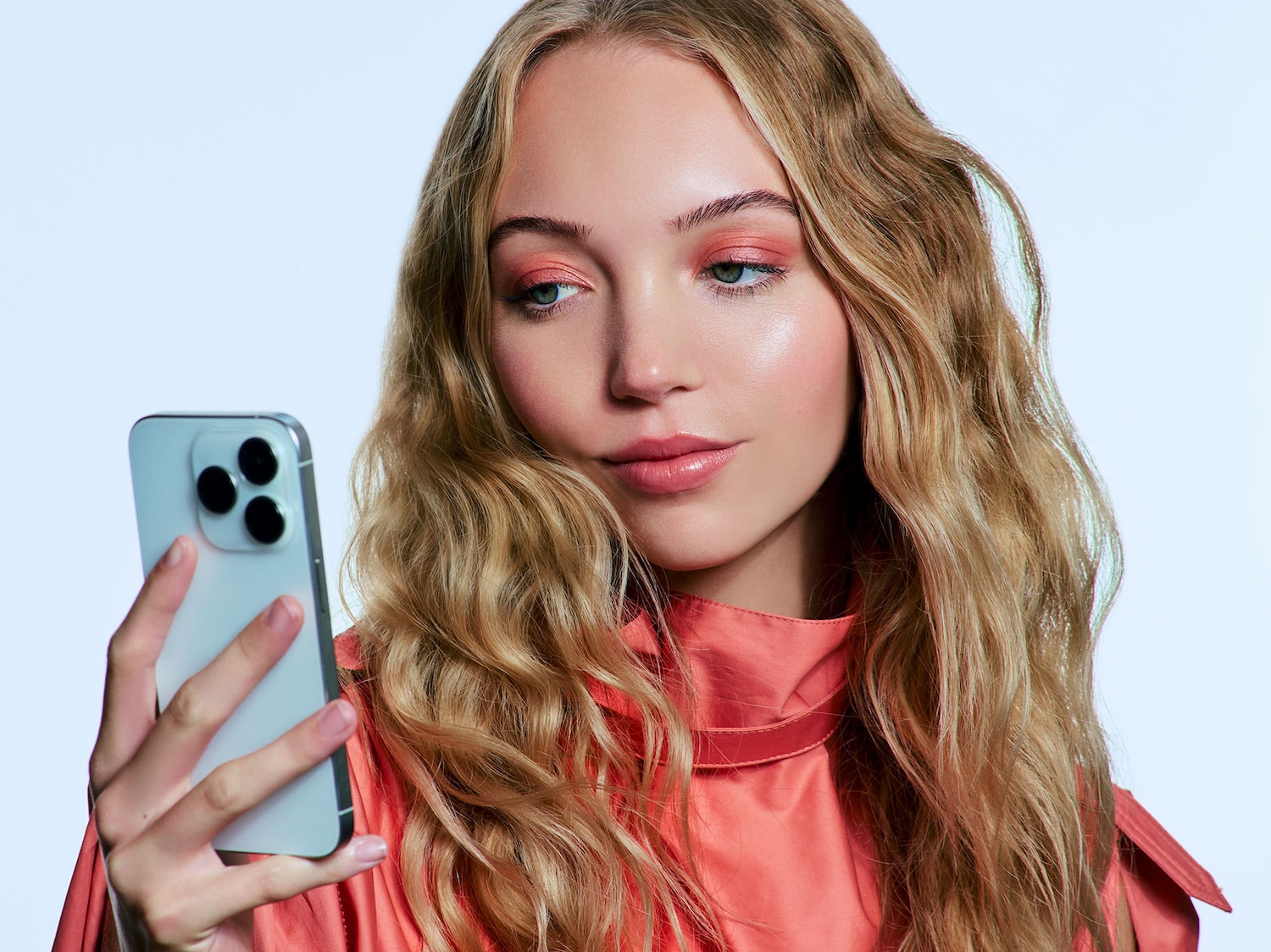 Primo piano di una donna bionda con ombretto e top color pesca che guarda il suo smartphone.