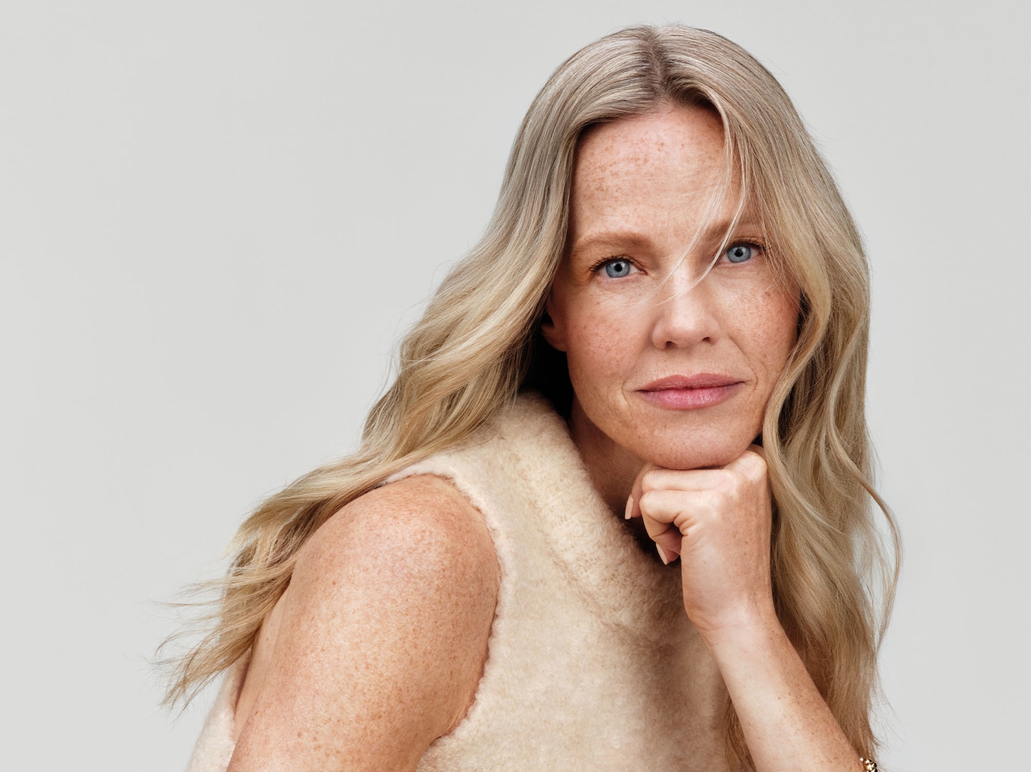 Primo piano di una donna bionda con gli occhi azzurri e la pelle chiara, appoggiata con il mento sulla mano, indossa un gilet di lana color crema.