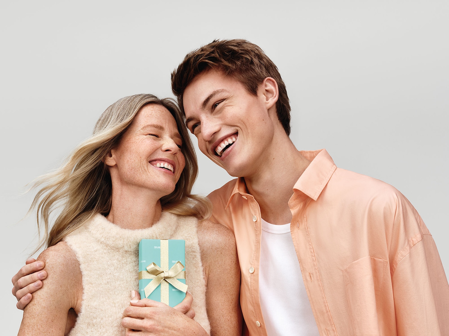 Una donna bionda e un giovane sorridono mentre lei tiene in mano un regalo di Douglas avvolto in carta azzurra e nastro dorato.