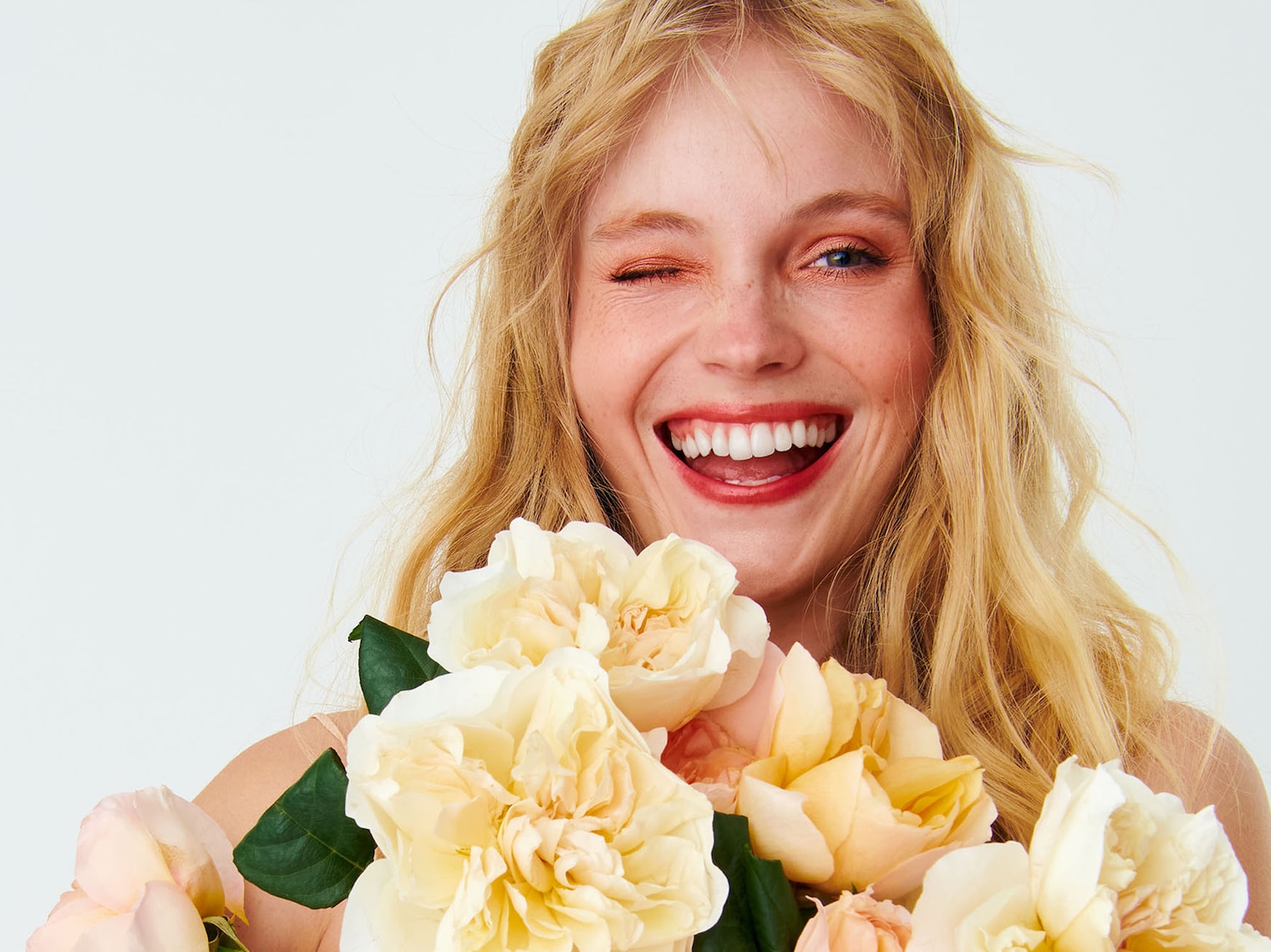 Una donna bionda sorride ammiccando mentre tiene in mano un bouquet di rose color crema e pesca, con un ombretto arancione brillante.