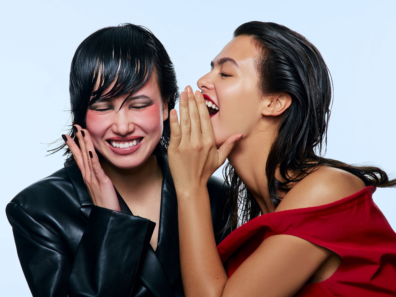 Due donne sorridono, una vestita di rosso che confida un segreto all'altra, con giacca di pelle nera.