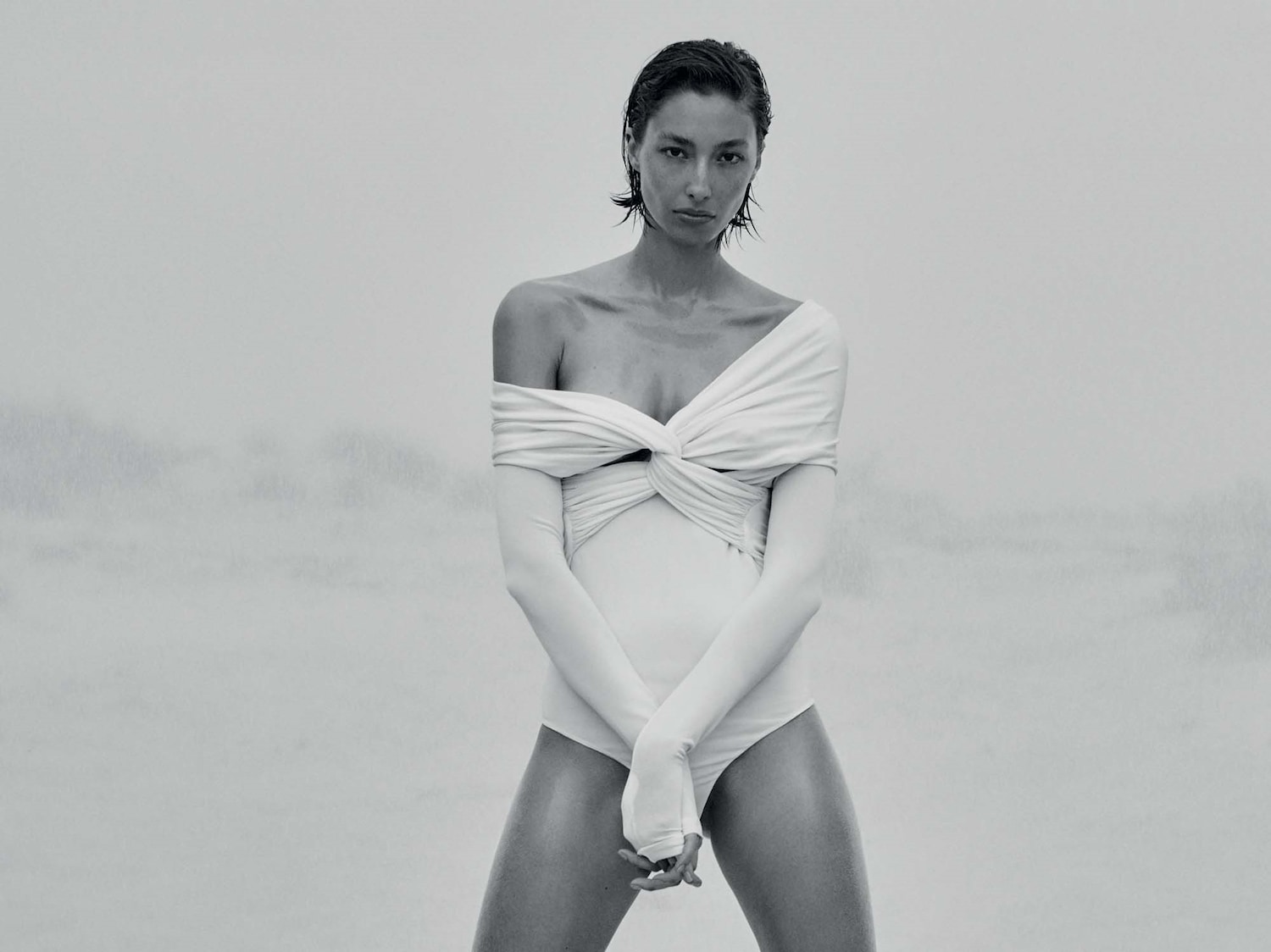 In uno scatto in bianco e nero, una modella con i capelli corti e bagnati indossa un costume intero bianco con maniche lunghe, posando con le braccia incrociate in un ambiente nebbioso.