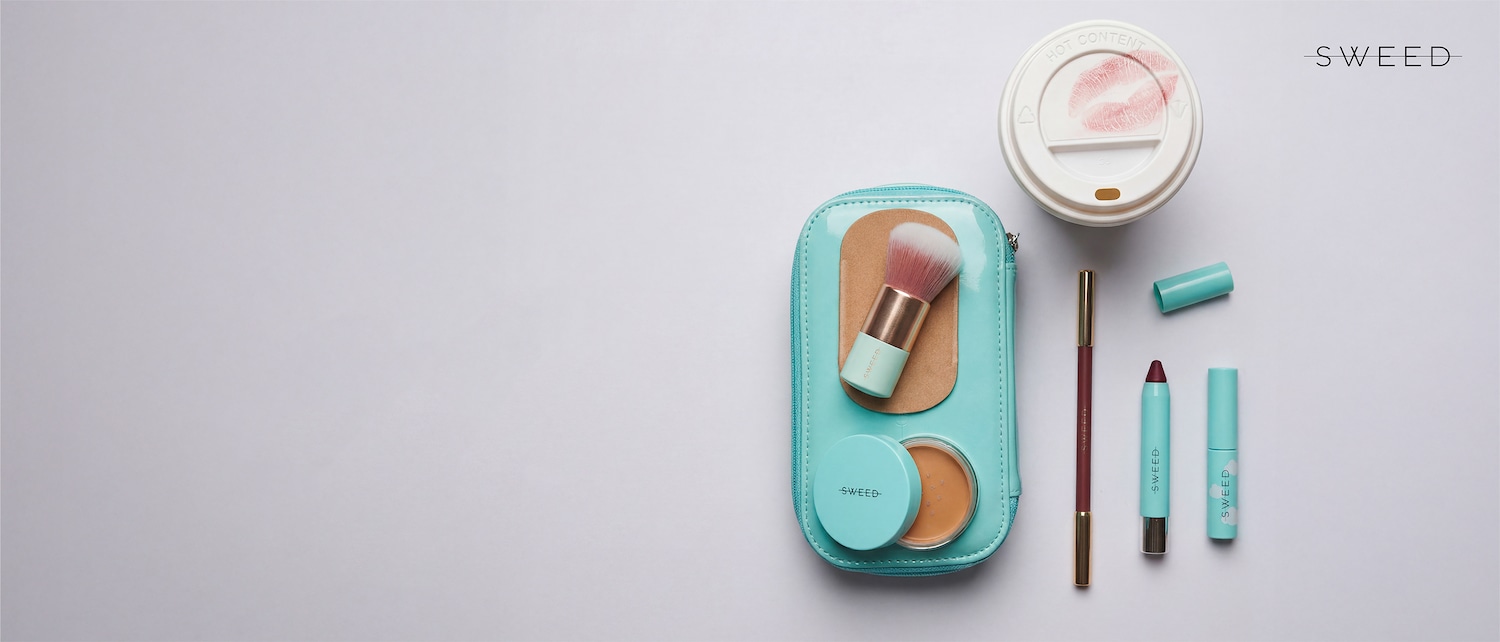 Una trousse azzurra lucida contiene un pennello da trucco con setole rosa e un fondotinta compatto color carne, accanto a una matita marrone, un rossetto bordeaux e un lucidalabbra, il tutto del marchio Sweed.