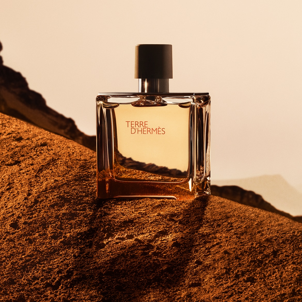 Il profumo Terre D'Hermès in una bottiglia di vetro trasparente squadrata con tappo nero, adagiata su un letto di terra marrone.
