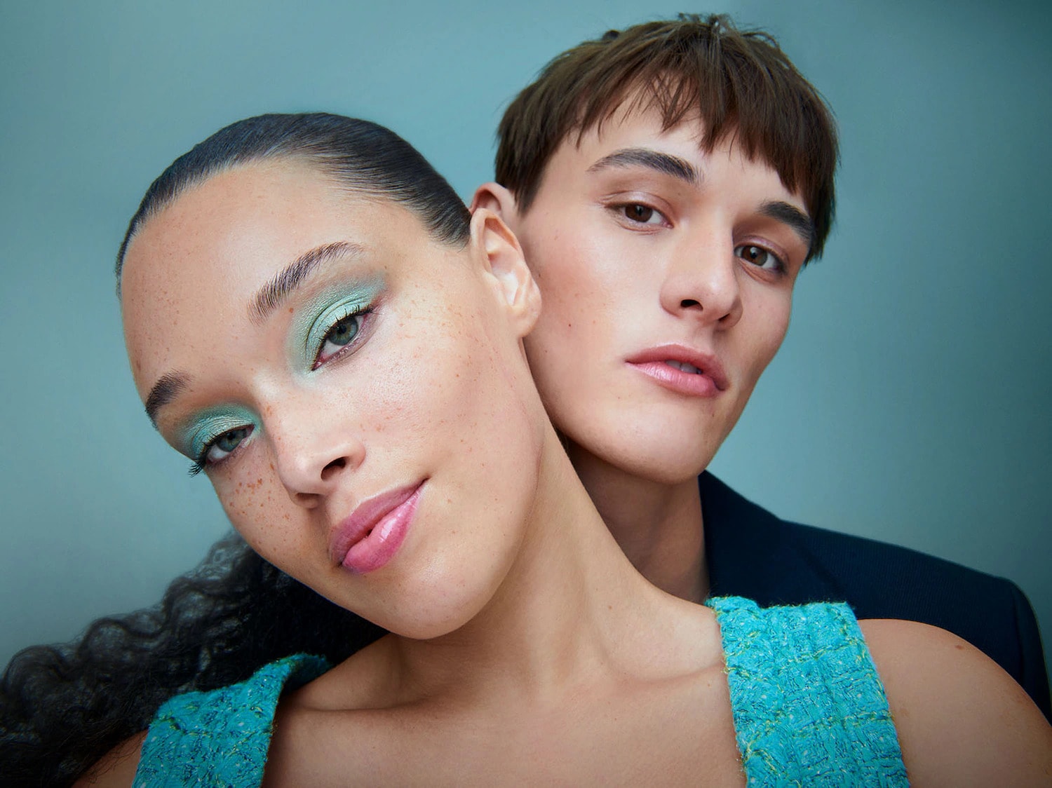 Una donna con ombretto verde acqua e labbra rosa lucide, con un uomo dietro di lei che indossa una giacca blu scuro, entrambi con un trucco naturale e fresco.