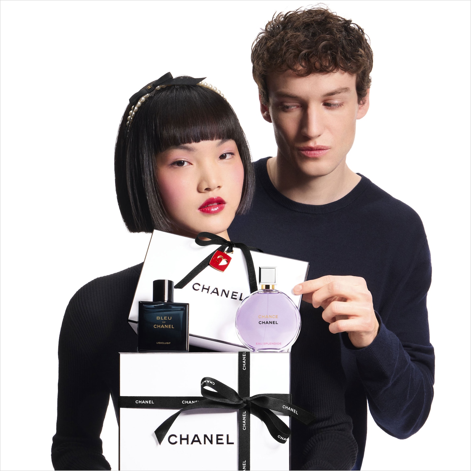 Una coppia presenta due confezioni regalo Chanel bianche, decorate con nastri neri logati, contenenti i profumi Bleu de Chanel e Chance Eau Splendide.