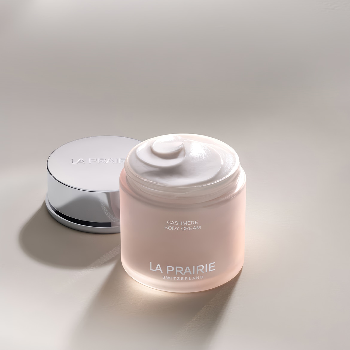 Ecco un barattolo aperto di crema corpo cashmere La Prairie, con il suo coperchio argentato appoggiato accanto, che mostra la consistenza ricca e vellutata della crema.