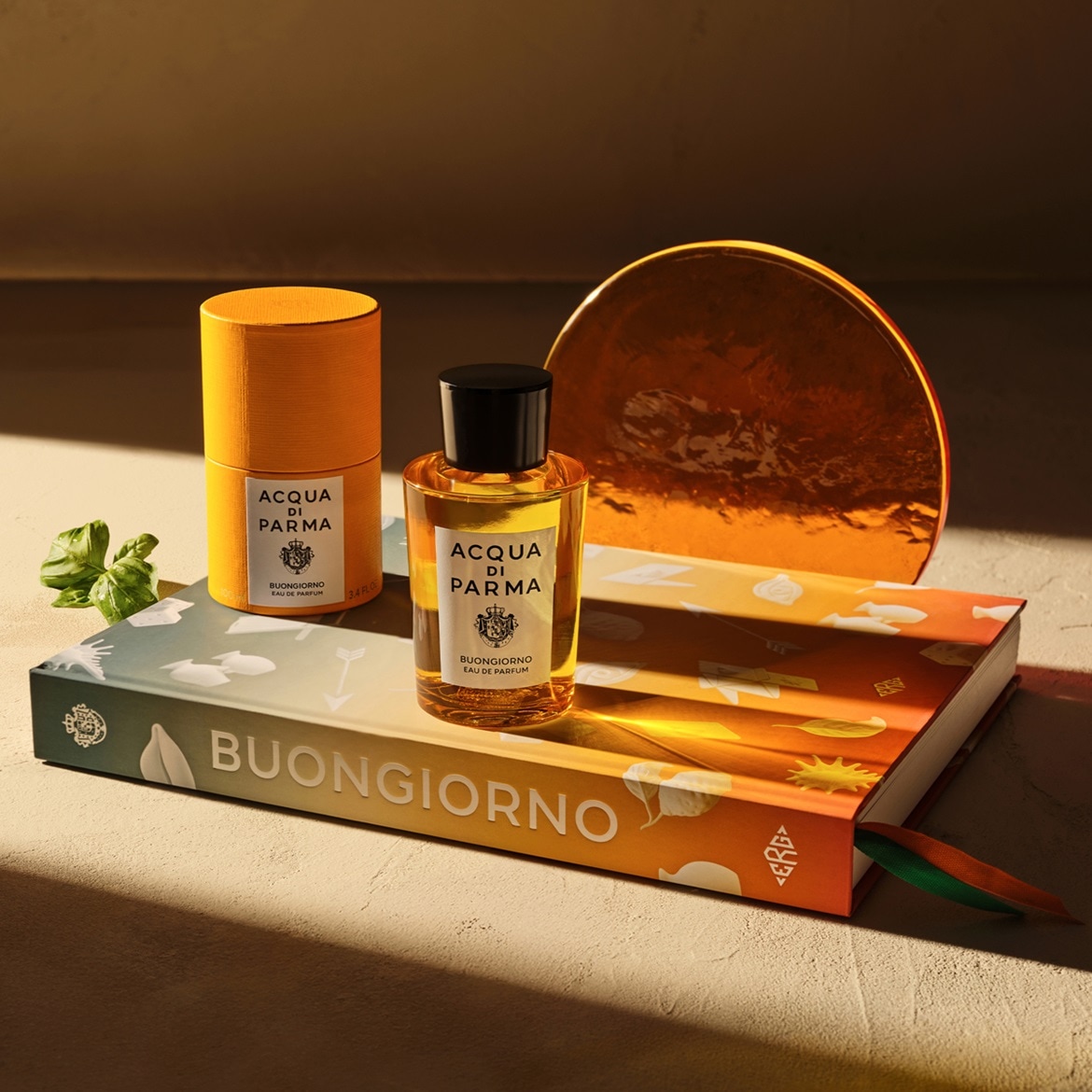 Il profumo Acqua di Parma Buongiorno Eau de Parfum in un flacone di vetro trasparente con tappo nero, accanto a una confezione cilindrica arancione, entrambi appoggiati su un libro colorato.