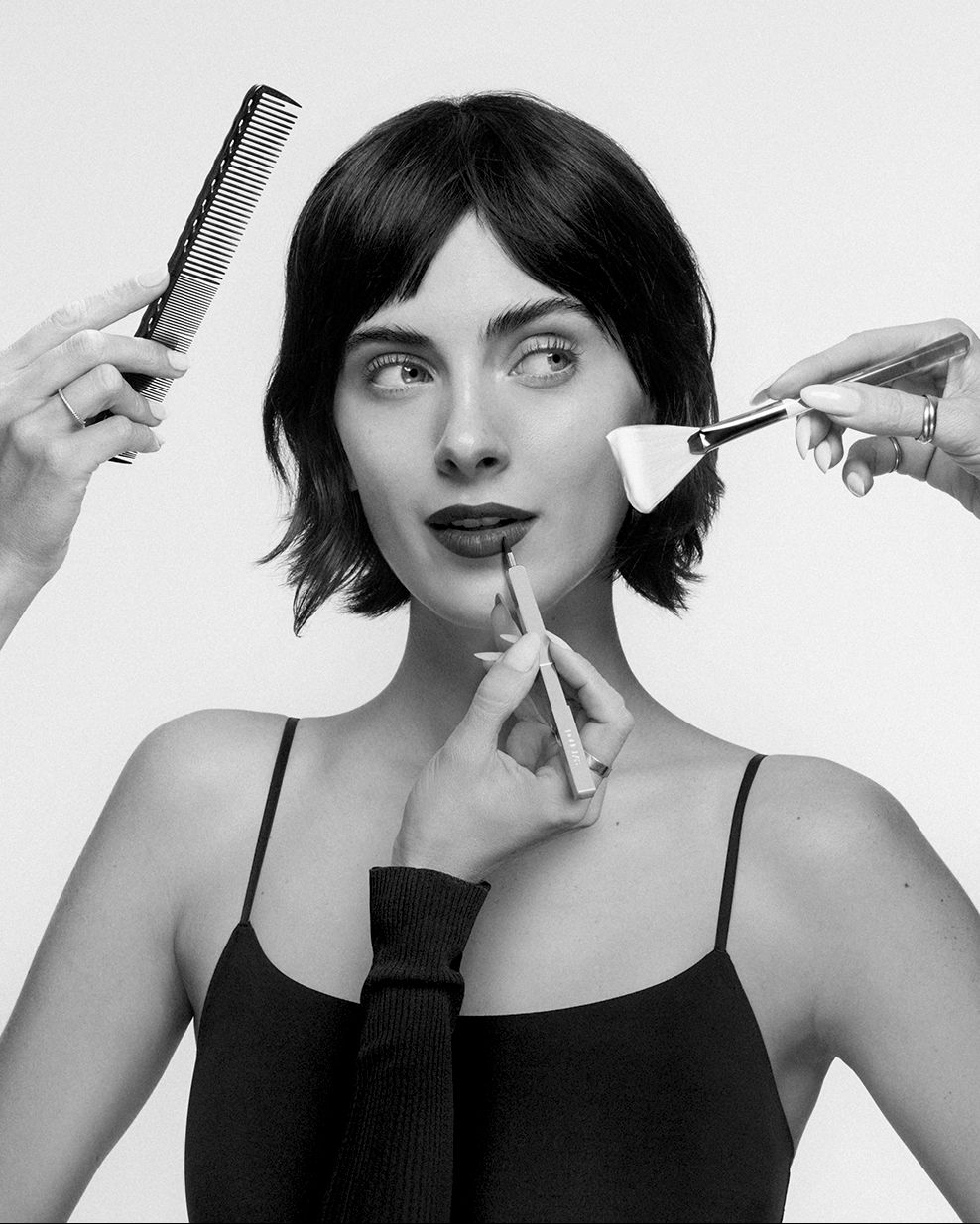 Una donna con capelli corti e scuri, indossa un top nero con spalline sottili, mentre le sue labbra sono delineate con una matita scura e un pennello per il viso applica un prodotto sulla guancia, mentre un pettine nero è tenuto vicino ai suoi capelli.